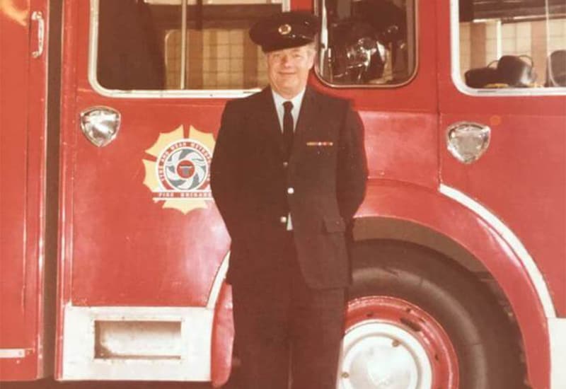 A man stands proudly in front of a vintage red fire truck, wearing formal firefighter uniform with insignia and a cap.
